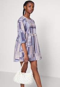 Vestido de manga curta, na altura dos joelhos, em padrões azuis e beges, com uma cintura franzida. A modelo segura uma bolsa de palha branca.