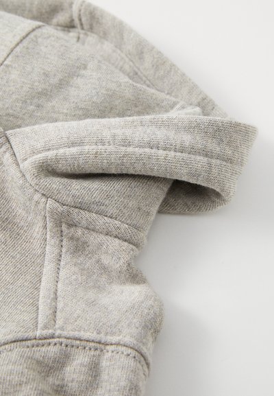 Lähikuva vaaleanharmaasta, taitellusta neulotusta kangassukkaasta, jossa näkyy saumat ja ompelun yksityiskohdat valkoisella taustalla.