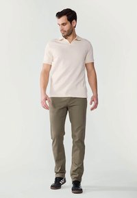 Άνδρας με μπεζ πουκάμισο κοντό μανίκι, ελαιοπράσινα παντελόνια και μαύρα αθλητικά παπούτσια στέκεται κοιτάζοντας χαμηλά αριστερά του σε λευκό φόντο.