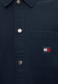 Σκούρο μπλε πουκάμισο με κουμπιά και γιακά, μεταλλικά κουμπιά με χαραγμένο "Tommy Jeans" και τσέπη στο στήθος με το έμβλημα Tommy Jeans.