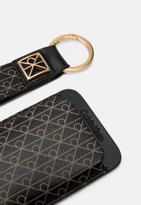 Coque de téléphone Calvin Klein en cuir noir, avec un motif géométrique doré et un anneau en métal doré pour l'attache. Détail de surface texturée.