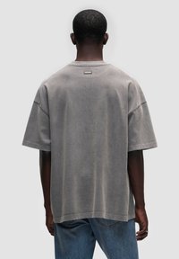 Przytulny, szary t-shirt z bawełny w rozmiarze oversized, z krótkimi rękawami i okrągłym dekoltem, z metką z tyłu. Noszony z niebieskimi dżinsami, co podkreśla swobodny styl.