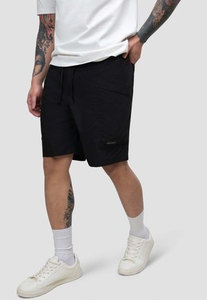 Mann trägt schwarze Shorts, weißes T-Shirt, weiße Socken und weiße Sneakers, zeigt Tätowierungen an Unterarmen und Unterschenkeln.