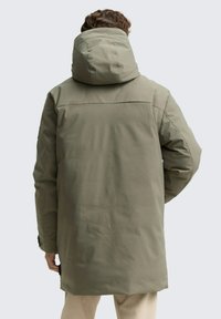 Olivno zelen park s visokim ovratnikom in snemljivo kapuco. Ima pravilen kroj, strukturno tkanino in stranske razpoke za lažje gibanje.