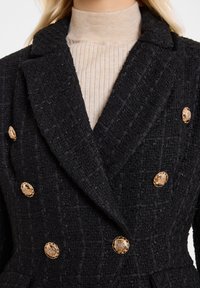 Zwarte gestructureerde blazer met goudkleurige decoratieve knopen, dubbele sluiting en brede revers, gedragen over een beige geribbelde turtleneck.