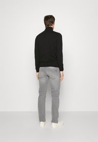 Svart, långärmad, höghalssweater i kombination med ljusgrå jeans och vita sneakers, sedd från baksidan mot en enkel bakgrund.