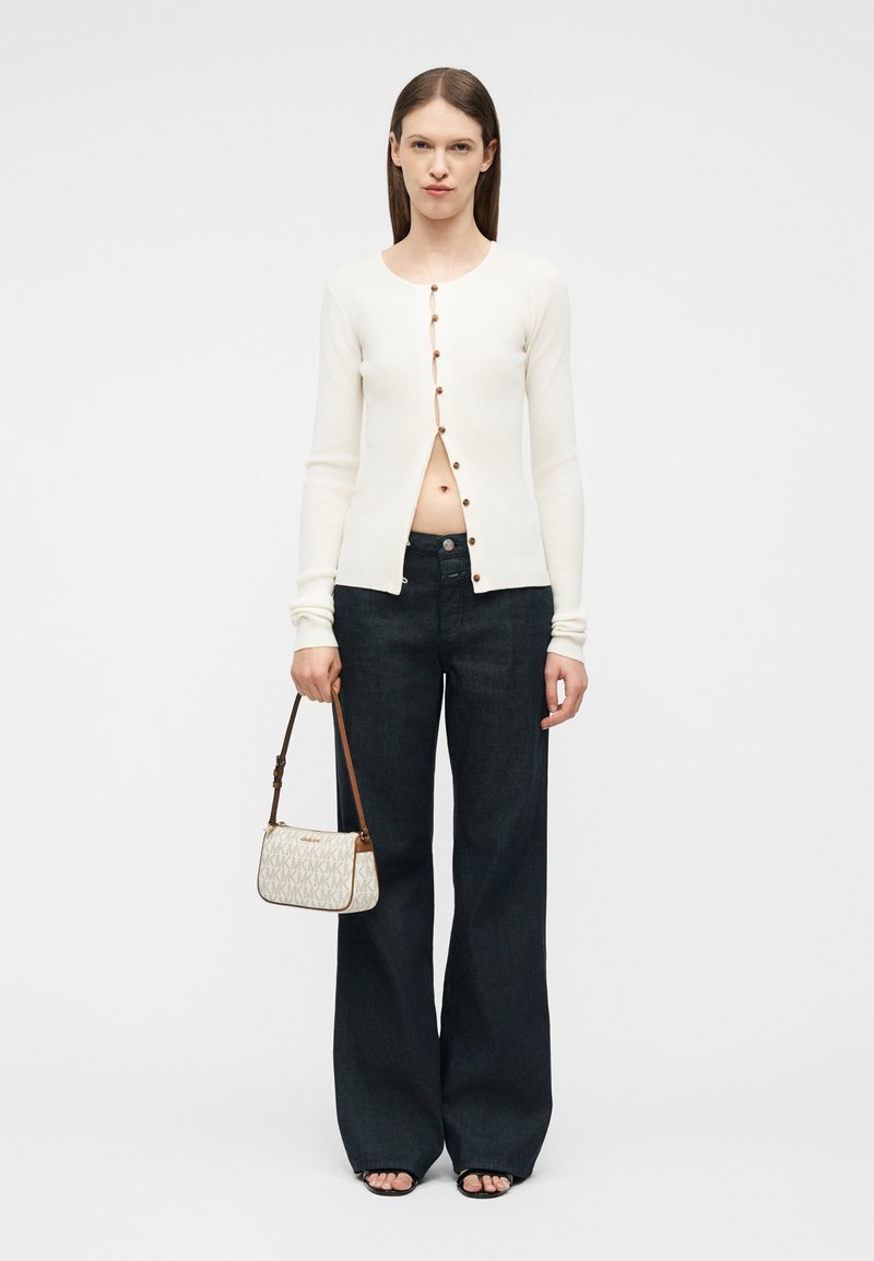 Femme portant un cardigan crème partiellement boutonné, un jean large sombre, des sandales noires, tenant un petit sac à main beige à motif.