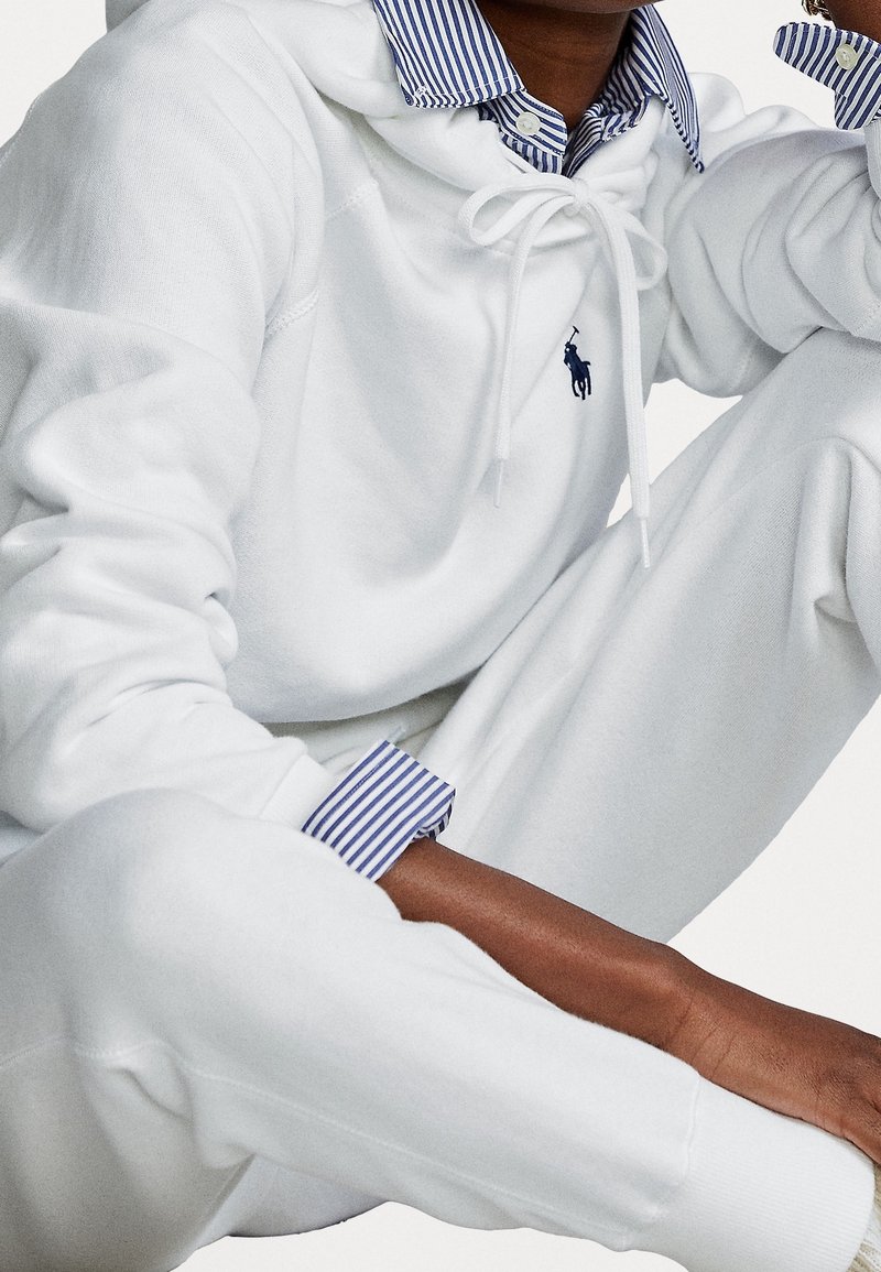 Personne en sweat à capuche blanc et pantalon de survêtement assise avec une chemise rayée bleue et blanche à col visible sous le sweat.