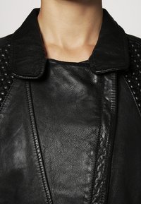 Giubbotto di pelle nera con colletto, caratterizzato da sezioni lisce e testurizzate, e cuciture dettagliate lungo la parte anteriore e le spalle.