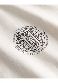 Logo s designem globusu, velké písmeno "H" uprostřed a text "LES DEUX COPENHAGEN PEČLIVĚ VYTVÁŘENO PRO LEPŠÍ ŽIVOT" na bílé tkanině.