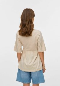 Bežs tops ar īsām piedurknēm un savāktu jostasvietu, saskaņots ar gaiši ziliem džinsu šortiem. Tops ir ar gludu, mīkstu tekstūru.