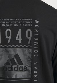 Musta t-paita, jossa on harmaalla printattu logo ja teksti, mukaan lukien "1949" ja "WORLDWIDE SPORT", asetettuna teksturoidun taustasuunnittelun päälle.