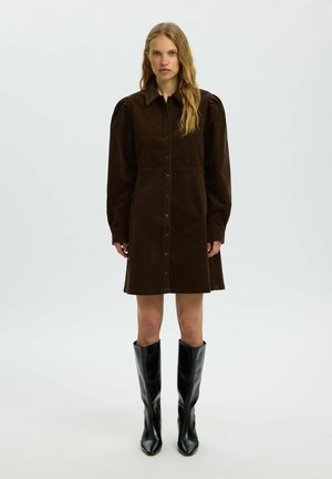 Femme aux longs cheveux blonds portant une robe boutonnée marron foncé avec des manches longues bouffantes et des bottes noires montant jusqu'aux genoux, debout devant un fond uni.