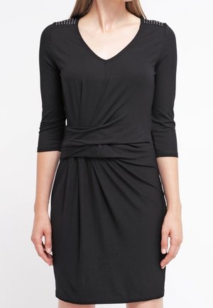 Woman wearing a black knee-length dress with three-quarter sleeves, V-neck, ruched waist, and stud details on the shoulders.