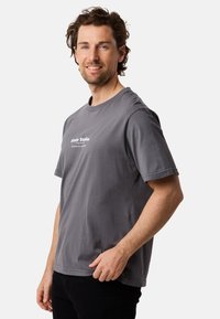 Harmaa puuvillainen t-paita, jossa on lyhyet hihat ja pyöreä kaula-aukko. Edessä valkoista tekstiä. Päällä mustat housut. Rentoa tyyliä.