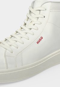 Λευκό sneaker ψηλής κορυφής με λευκά κορδόνια, κόκκινο λογότυπο "HUGO" στο πλάι και λεπτομερείς ραφές σε λείο δερμάτινο υλικό.