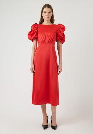 Mujer de pie que lleva un vestido midi rojo con mangas cortas abullonadas y tacones negros de punta afilada, sobre un fondo claro y liso.