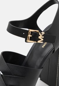 Sandale negre cu toc înalt și platformă groasă, având curele intersectate și un accent cu cataramă aurie. Textură de piele netedă și design elegant.