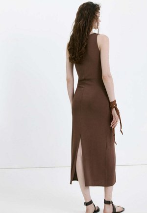 Mujer con el cabello largo y rizado que lleva un vestido midi marrón sin mangas con abertura trasera, sandalias negras con tira en el tobillo y un pañuelo marrón en la muñeca.