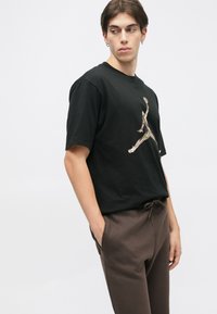 Schwarzes Baumwoll-T-Shirt mit kurzen Ärmeln, das auf der Vorderseite ein kamouflage-musterndes Jumpman-Logo zeigt. Das Model trägt braune Sweatpants mit Kordelzug.