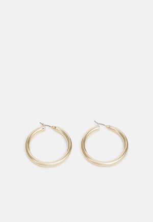 Zlaté středně velké kruhové náušnice s tenkými kovovými tyčinkami na jednoduchém světle šedém pozadí.