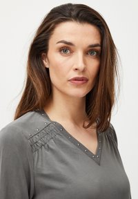 Hall Gray blouse koos V-kaelusega, millel on kogutud detailid ja dekoratiivsed hõbedased nupukesed õlgadel. Sile kangas.