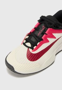 Športová obuv s bielym textúrovaným zvrškom, čiernymi šnúrkami, ružovými akcentmi a výrazným červeným logom Nike. Obsahuje čiernu gumovú podrážku.