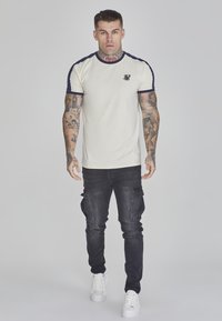 Camiseta de manga corta cremosa con detalles en azul marino, que presenta un pequeño logo en el pecho, combinada con pantalones cargo oscuros y zapatillas blancas.