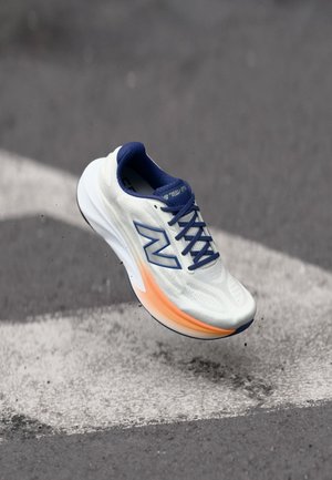 Weißer und blauer Laufschuh mit orangefarbenem Sohlenakzent, in der Luft über einer Asphaltstraße mit weißen Fahrbahnmarkierungen.