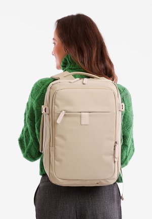 Beige Rucksack mit mehreren Fächern, glatter Textur und Reißverschluss-Verschlüssen. Über einer Schulter getragen, kombiniert mit einem grünen Strickpullover.