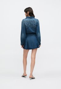 Džínsové šaty s dlhými rukávmi, priliehavým pásom a rozšírenou sukňou. Na zadnej strane dekoratívne prešitie. Modrá farba s opotrebovanými detailmi.