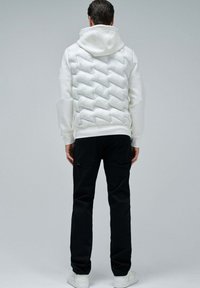 White puffer vest with a quilted design, paired with black pants and white sneakers, viewed from the back. Hooded neckline noted.
