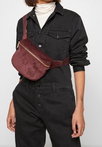 Osoba w czarnej dżinsowej kurtce i spodniach, z białym golfem, nosząca bordową torbę na biodra z sztruksu przerzuconą przez pierś.