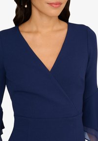 Robe bleu marine avec un décolleté en V profond, un design enveloppant et des manches évasées avec des accents transparents. Fabriquée dans un tissu lisse avec une coupe ajustée.
