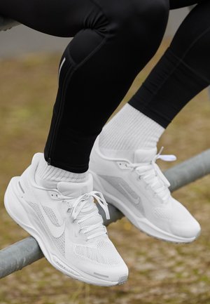 Persoon draagt witte sneakers en witte sokken, zittend op een metalen reling met gekruiste benen, gekleed in zwarte sportbroek.
