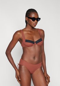Rjav bikini s strukturirano zgornjim delom, ki ima črne detajle. Prilagodljive stranske vezalke na dnu. Gladka tekstura, minimalističen dizajn.