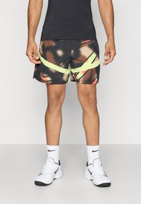 Pantalones cortos negros con patrones abstractos en naranja y amarillo, con una franja verde neón. Llevados con una camiseta negra y zapatillas deportivas blancas.