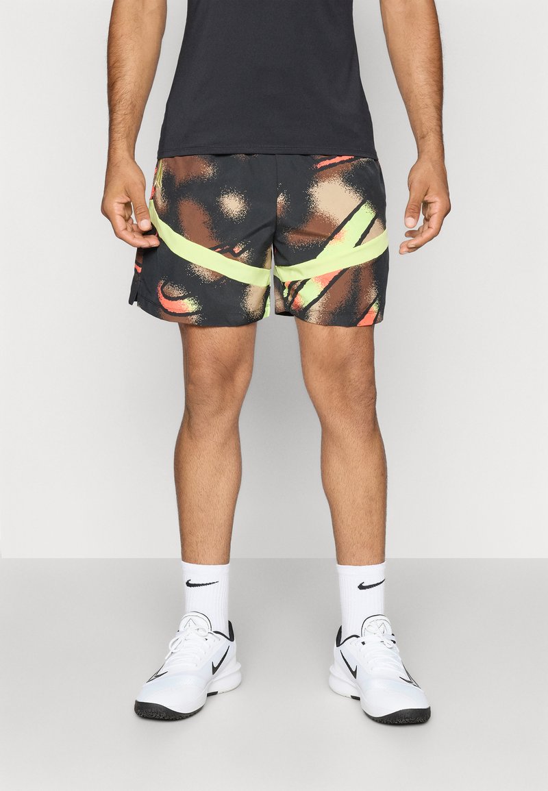 Pantalones cortos negros con patrones abstractos en naranja y amarillo, con una franja verde neón. Llevados con una camiseta negra y zapatillas deportivas blancas.