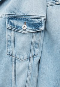 Poche de veste en denim bleu clair avec rabat sécurisé par un bouton métallique marqué "HUGO JEANS", comportant des coutures orange détaillées.