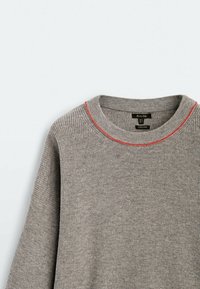 Pull gris, texture côtelée, encolure ronde avec accent rouge, manches longues, tissu doux, étiquette visible sous le col.