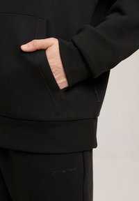 Czarna bluza z kangurzą kieszenią, ściągaczami przy mankietach i u dołu, miękka w dotyku, z dyskretnym logo na dole.