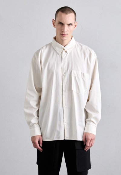 Chemise à manches longues couleur crème en tissu doux, avec un col à boutons et une poche poitrine, de coupe décontractée.