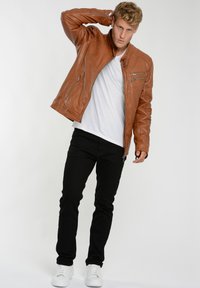 Brun læderjakke med høj krave og lynlåsdetaljer, parret med en hvid t-shirt, sorte bukser og hvide sneakers.