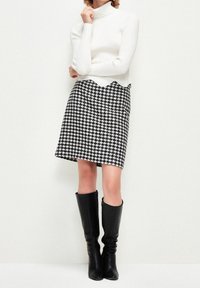 Col roulé en tricot couleur crème et jupe pied-de-poule noir et blanc, assortis à des bottes noires brillantes montantes jusqu'aux genoux, présentés sur un fond neutre.