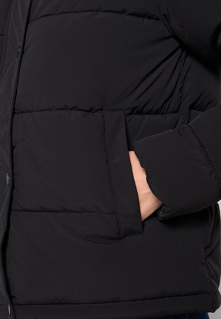 Doudoune noire avec un design matelassé, un col montant et une poche latérale, dotée d'une texture douce et d'une finition légèrement brillante.