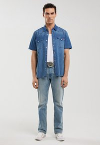 Camicia di jeans a maniche corte di colore azzurro chiaro, con colletto a punta, bottoni a scatto e due tasche sul petto, abbinata a jeans blu sbiaditi e sneakers bianche.