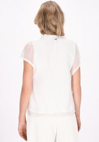 Weiße durchsichtige Bluse mit kurzen Ärmeln, Rüschensaum, strukturierten Polka-Dots und gewelltem Saum. Leichter Stoff, weiche Haptik.