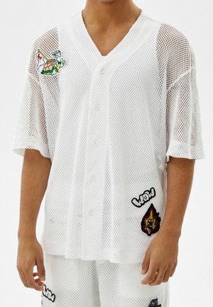 Homme portant un ensemble chemise à boutons en maille blanche et short avec des patchs de dessins animés, dont une tortue, une flamme et le texte « wow ».