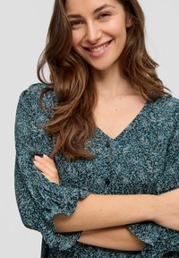 V-Ausschnitt-Bluse mit Struktur in blau-schwarzem Muster, ausgestattet mit drei Knöpfen und Rüschensleeves. Model mit verschränkten Armen, lächelt.