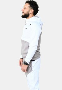 Sudadera con capucha gris y blanca de textura suave, con un logo en el pecho y un bolsillo canguro. Combinada con pantalones a juego.